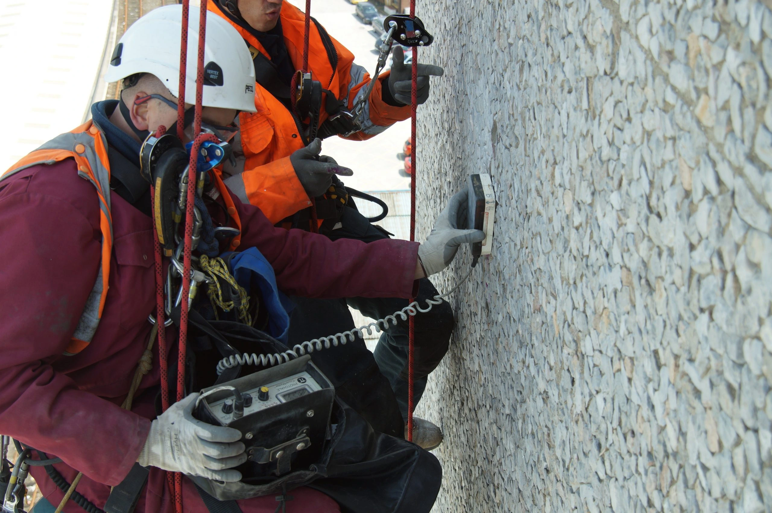 wo rope access technicians conducting a structural inspection on a high-rise building facade using specialist testing equipment to assess wall integrity.