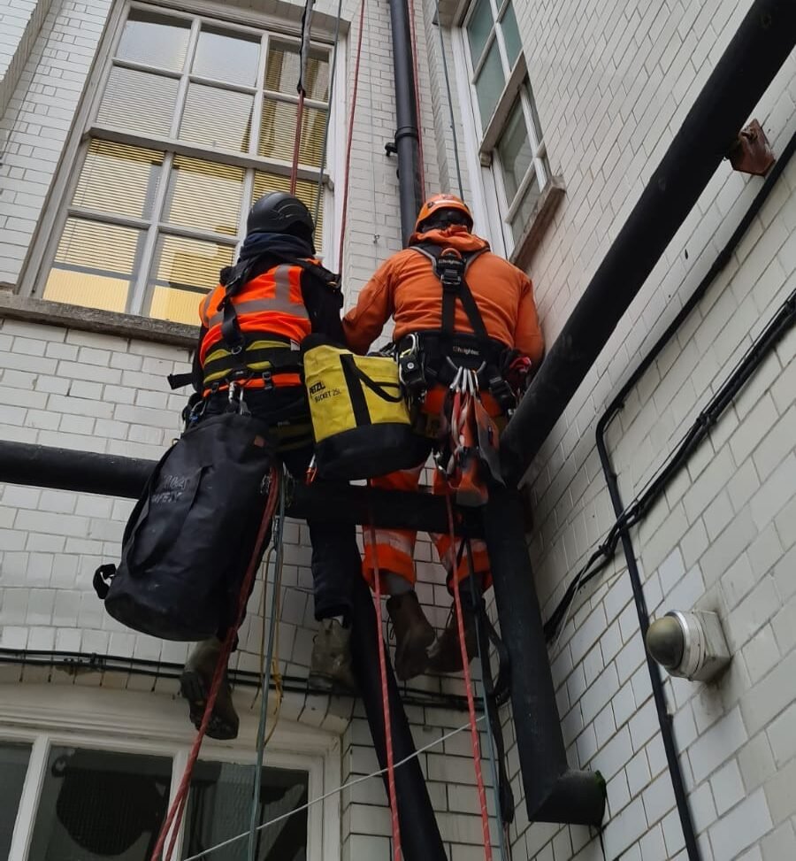 Rope access technicians inspecting and maintaining down pipe drainage on a high rise building in London to ensure proper water flow and prevent blockages