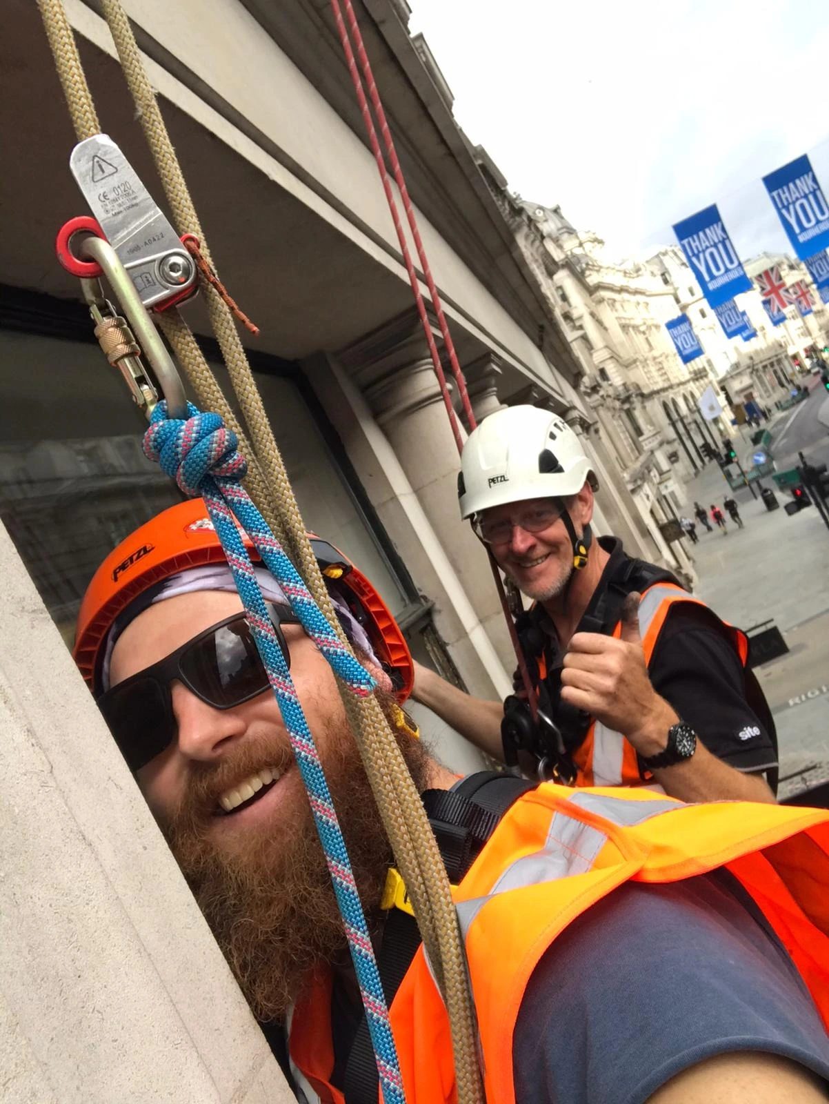 Two rope access technicians wearing safety gear and harnesses while working at height on a building facade in London. One technician is smiling at the camera, while the other gives a thumbs-up. Ropes and safety equipment are securely rigged
