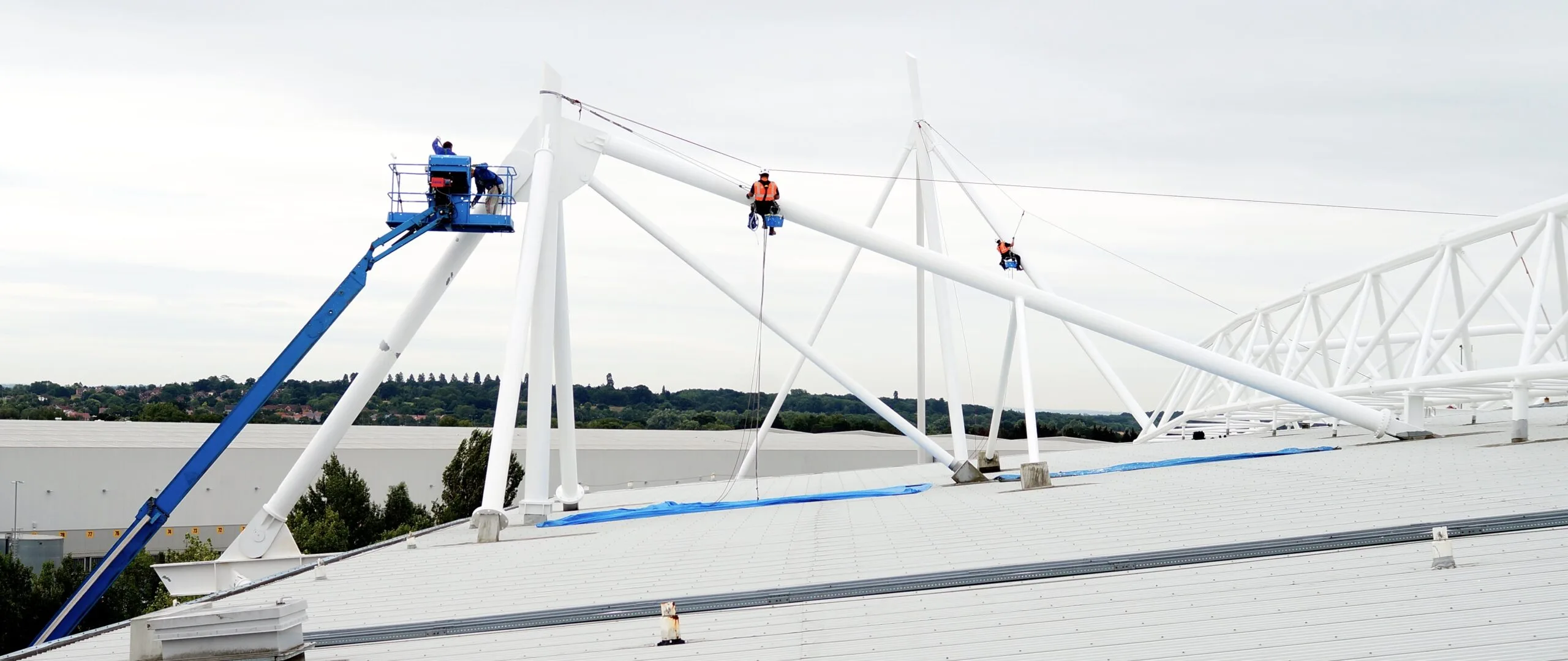 Rope access technicians and MEWP operator working on structural steel beams above a large roof